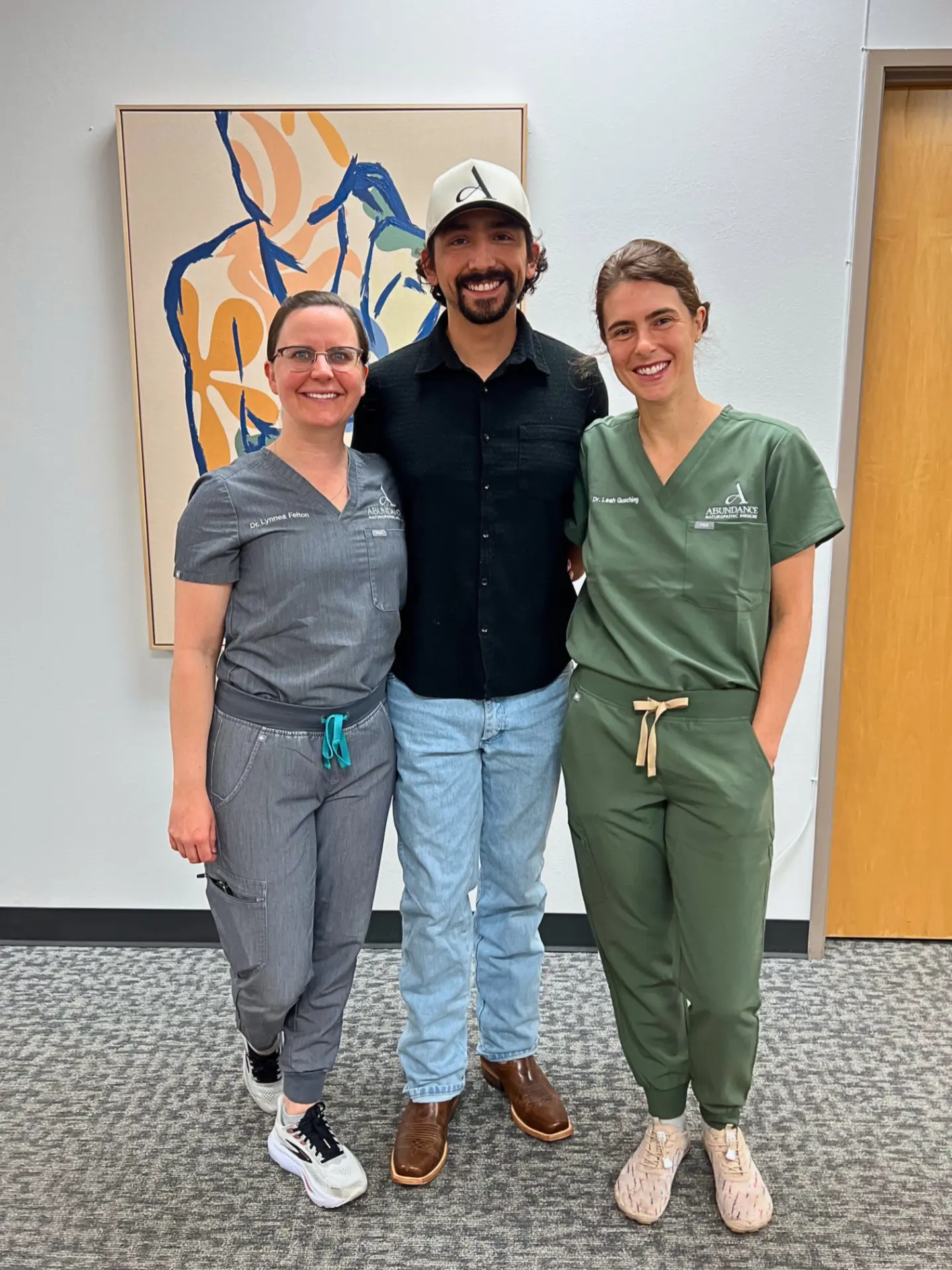 Dr. Lynnea Felton and Dr. Leah Gusching in scrubs smiling beside a guest in an Abundance cap